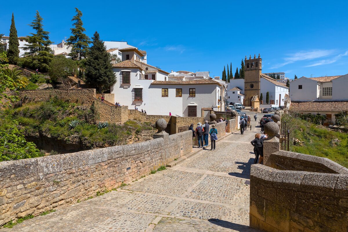 The old stone Puente Viejo bridge in Ronda leading to white houses and a church tower, with walkers on the bridge