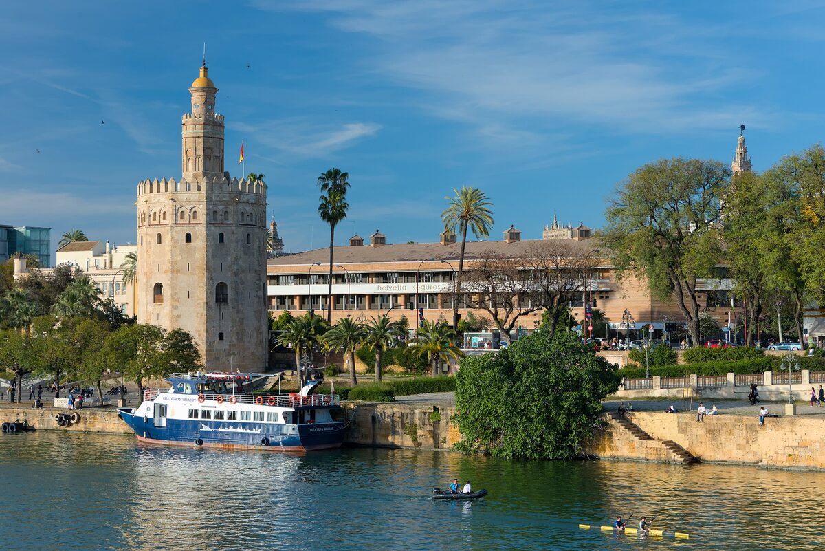 The twelve-sided Torre del Oro on the bank of the Guadalquivir, with a tourist boat on the water and palm trees along the river