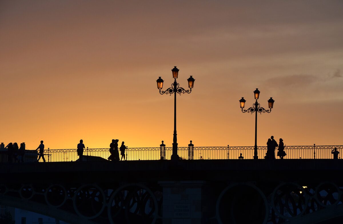 People walking across the Triana Bridge at sunset, silhouetted against an orange sky with old street lamps