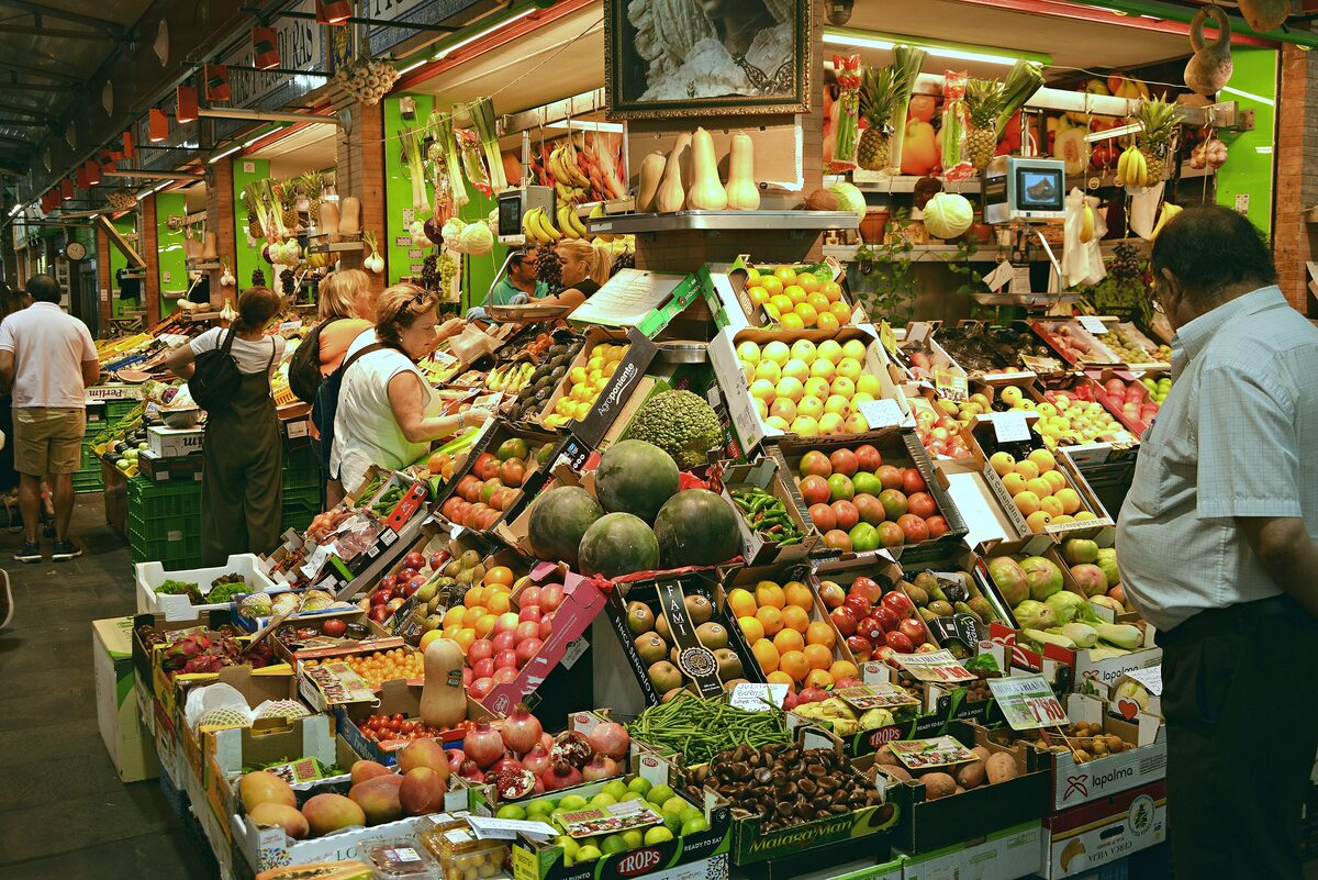 A fruit and vegetable stall piled with produce inside the Mercado de Triana, with shoppers at the counter