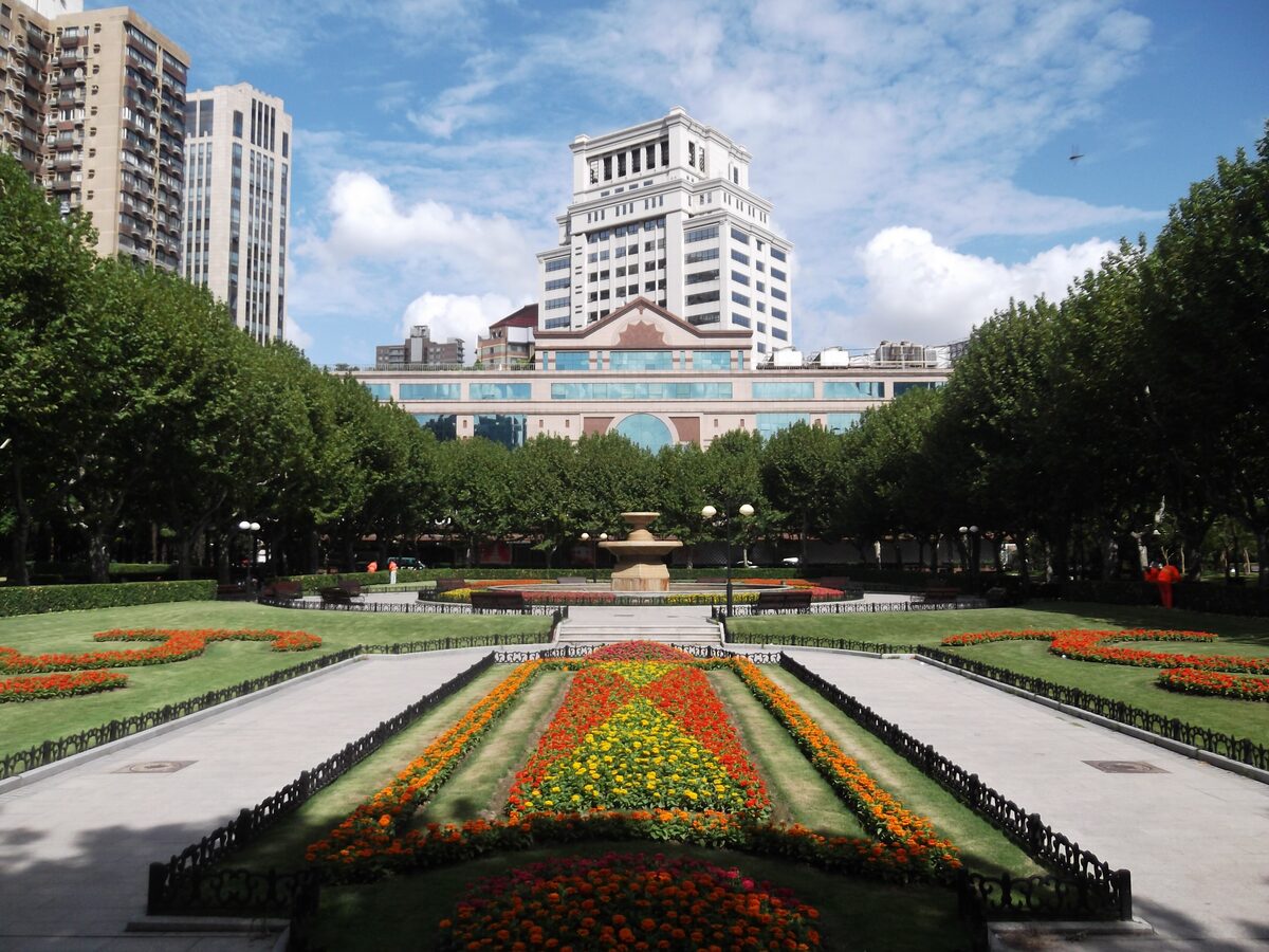 Formal flower beds and tree-lined lawns in Fuxing Park, with a white apartment tower behind the trees