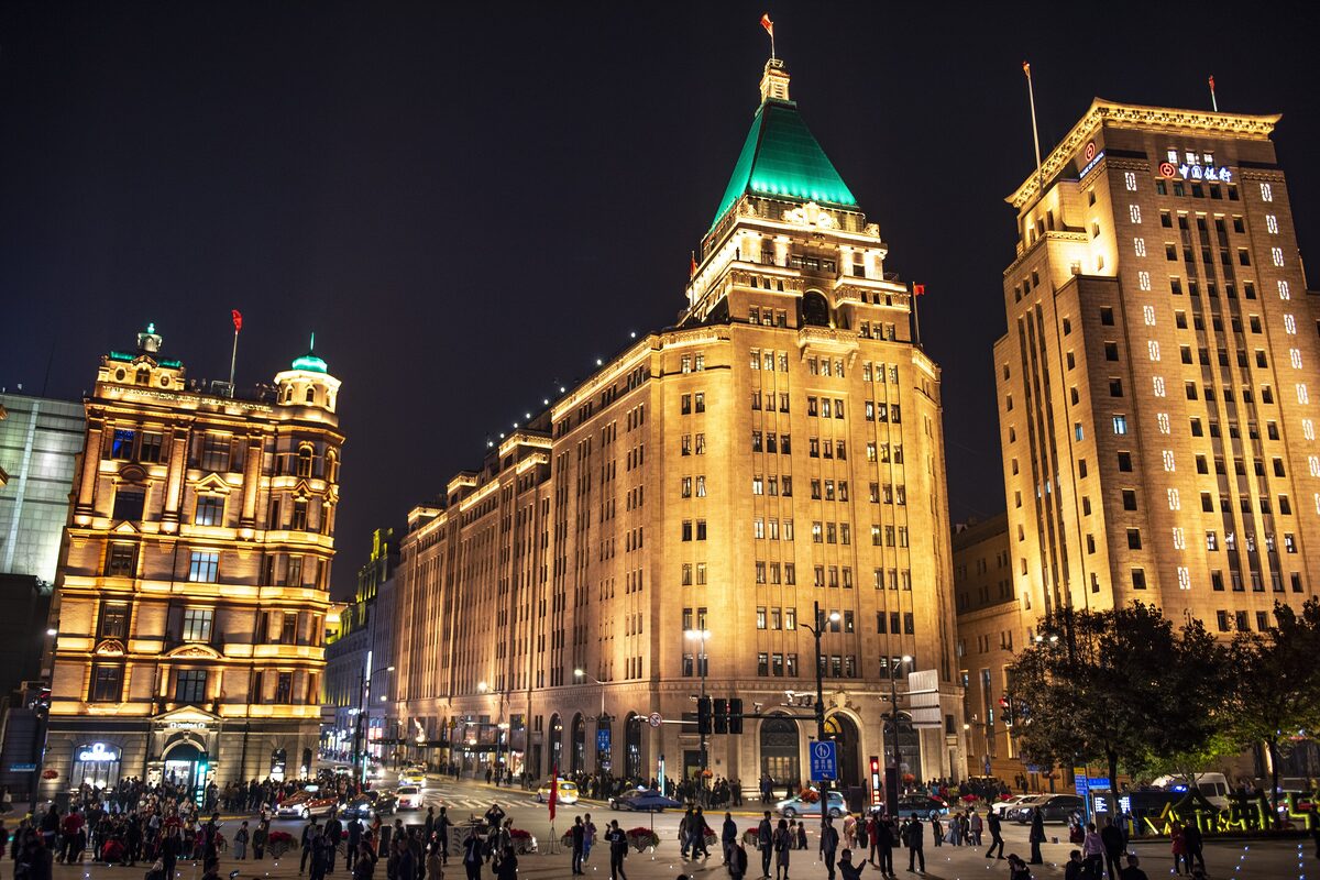 The green pyramid roof of the Fairmont Peace Hotel and the neighboring buildings lit up at night, with crowds on the street