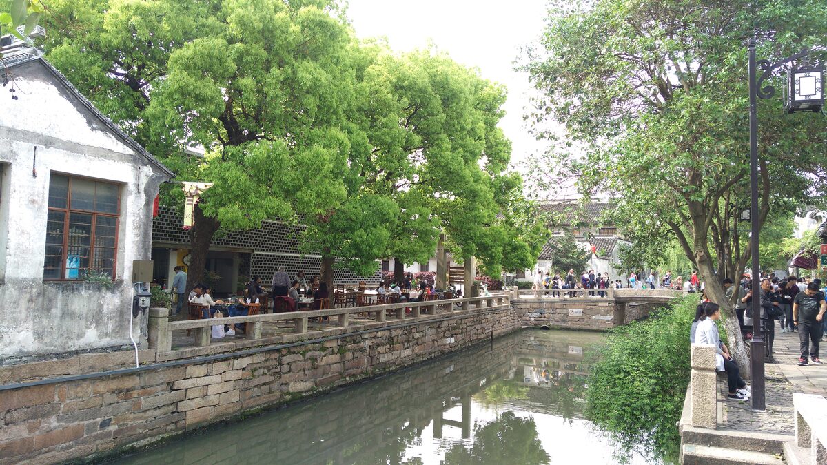 The Pingjiang canal with a stone embankment and shady trees, with people sitting along the water