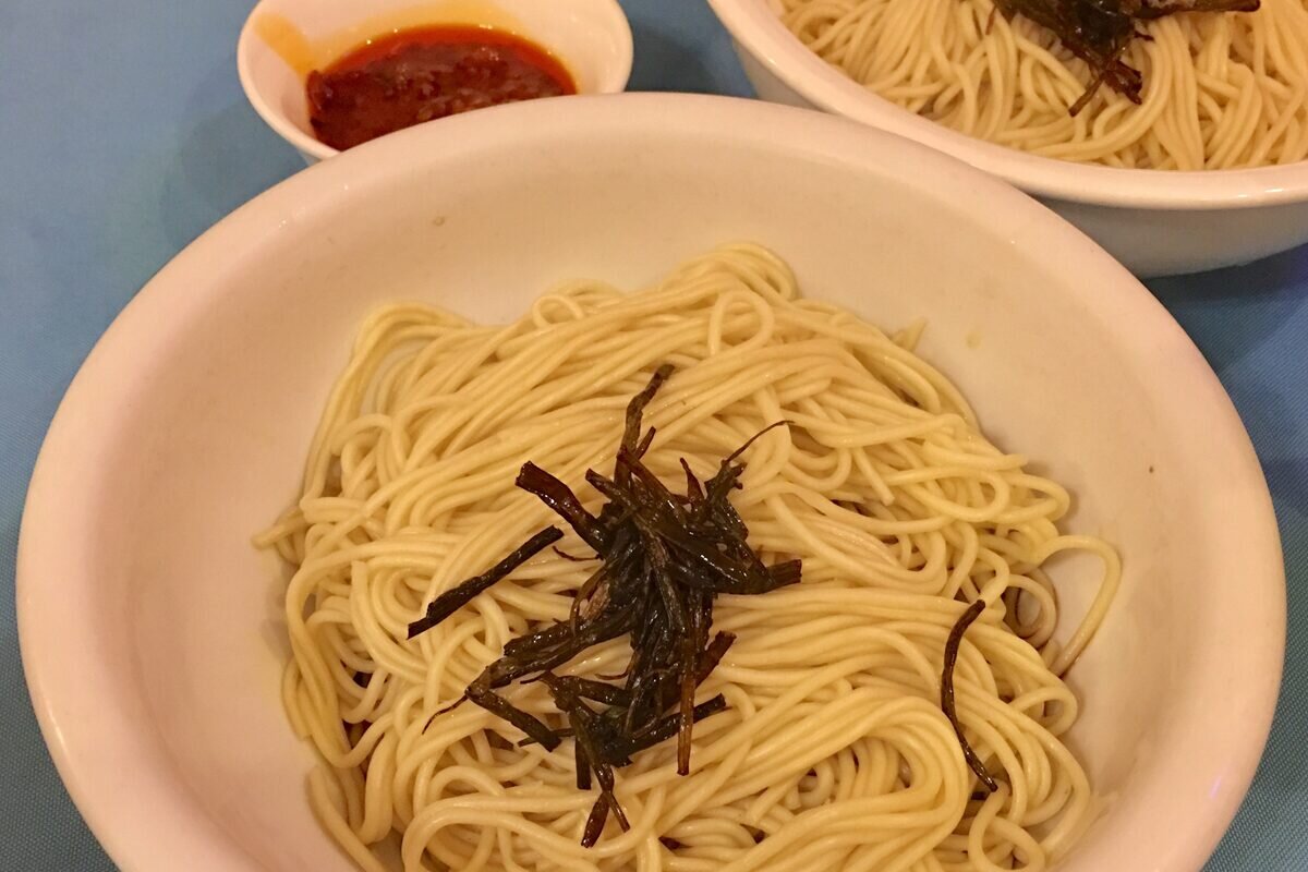 A bowl of plain noodles topped with crisp fried scallion, with a dish of chili sauce beside it