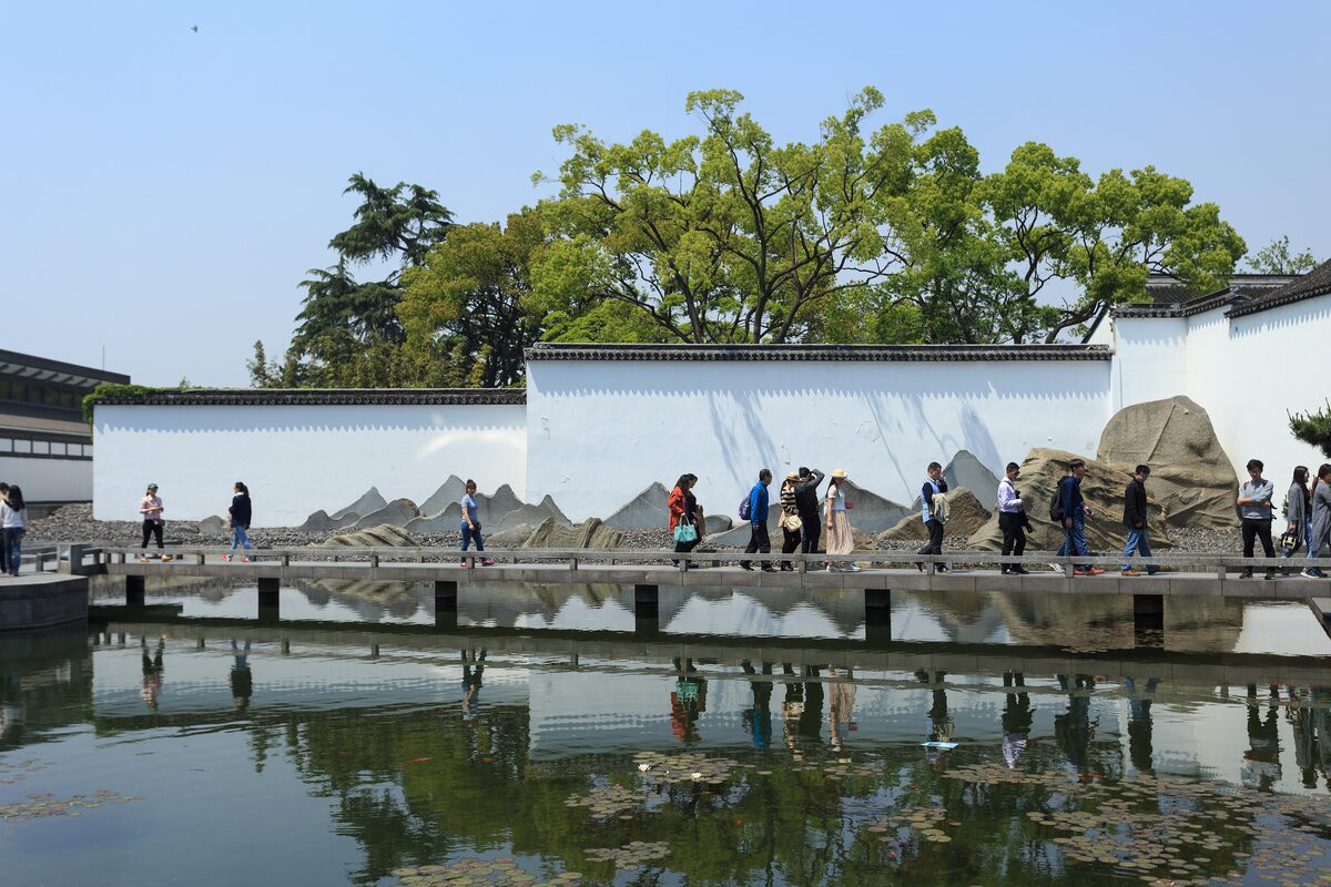 Visitors on a walkway beside the pond, with the white garden wall and the stone landscape in front of it at the Suzhou Museum
