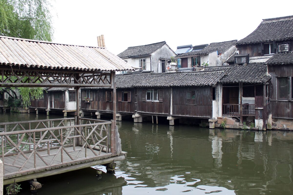 Dark-timber houses on stilts over the canal in Wuzhen, with a bamboo-roofed wooden jetty and a willow in the foreground