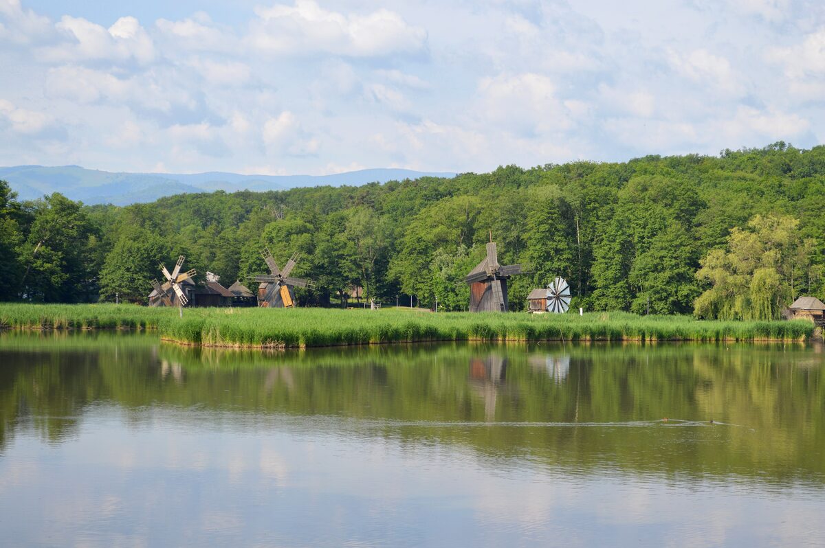 Wooden windmills and old farm buildings on the far shore of a calm lake at the ASTRA open-air museum, with forested hills behind