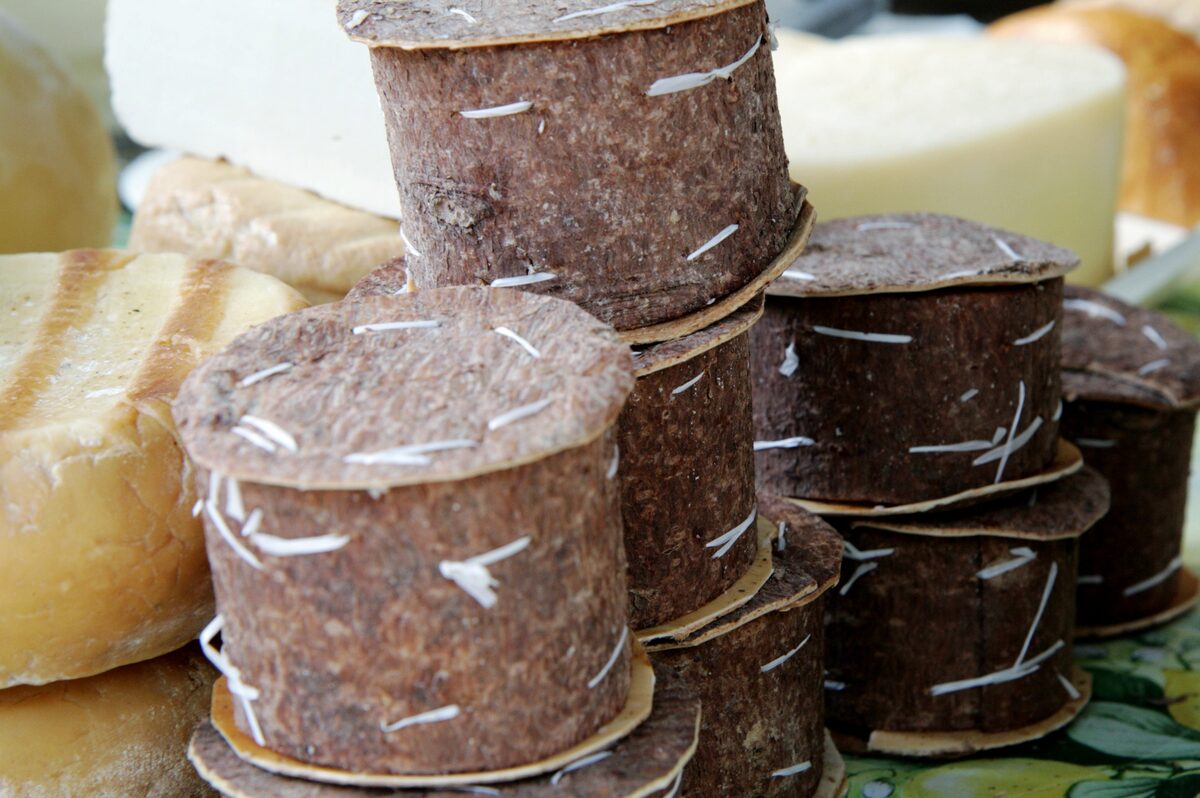 Stacks of brânză de burduf packed in fir-bark rolls on a market stall, next to rounds of smoked and white cheese
