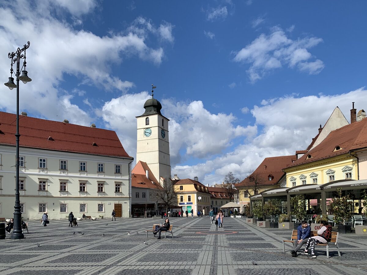 Piața Mare's patterned paving under a blue sky, with the Council Tower and colorful burgher houses with café terraces