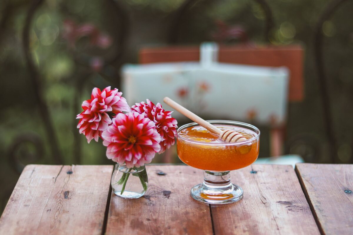 A glass dish of golden honey with a wooden dipper on a table, with a small vase of pink dahlias beside it