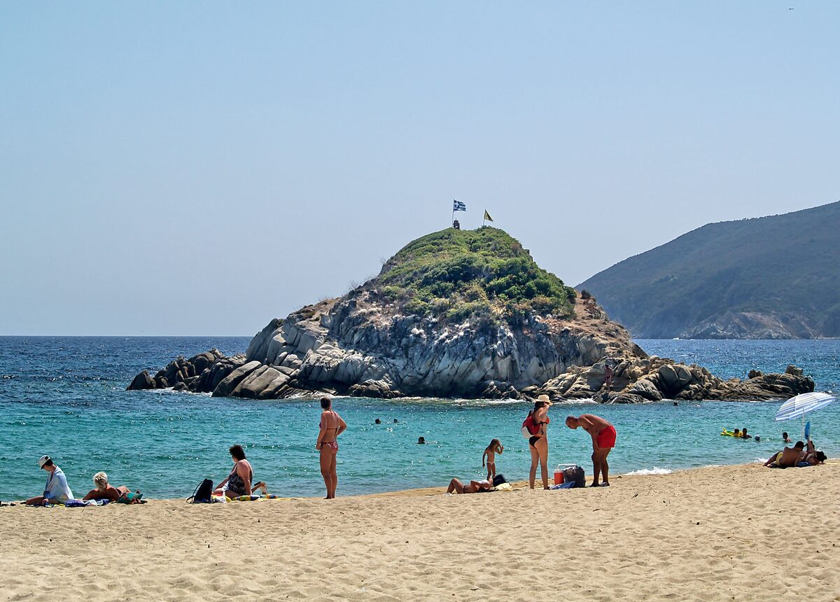 A small rocky islet with two flags rising off the sand at Kalamitsi beach, with bathers on the shore