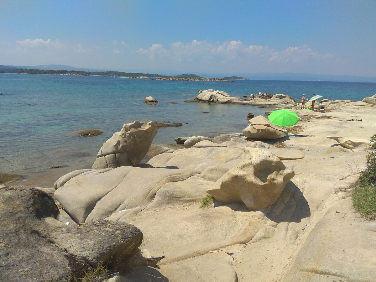 Sculpted pale granite boulders at Karidi beach beside clear turquoise water, with a green umbrella and sunbathers on the rocks