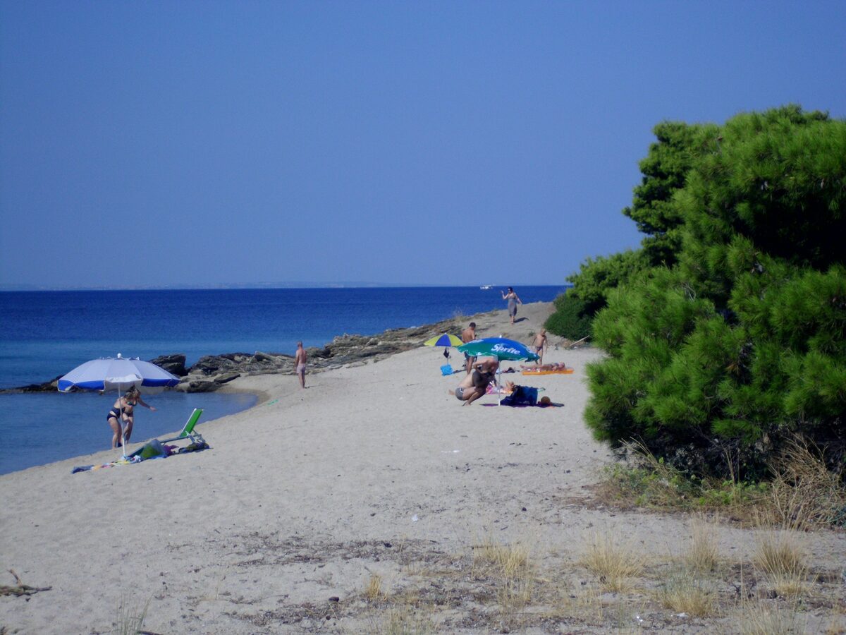 Sun umbrellas and bathers on the sand of Lagomandra beach, with pines and a rocky point along the shore