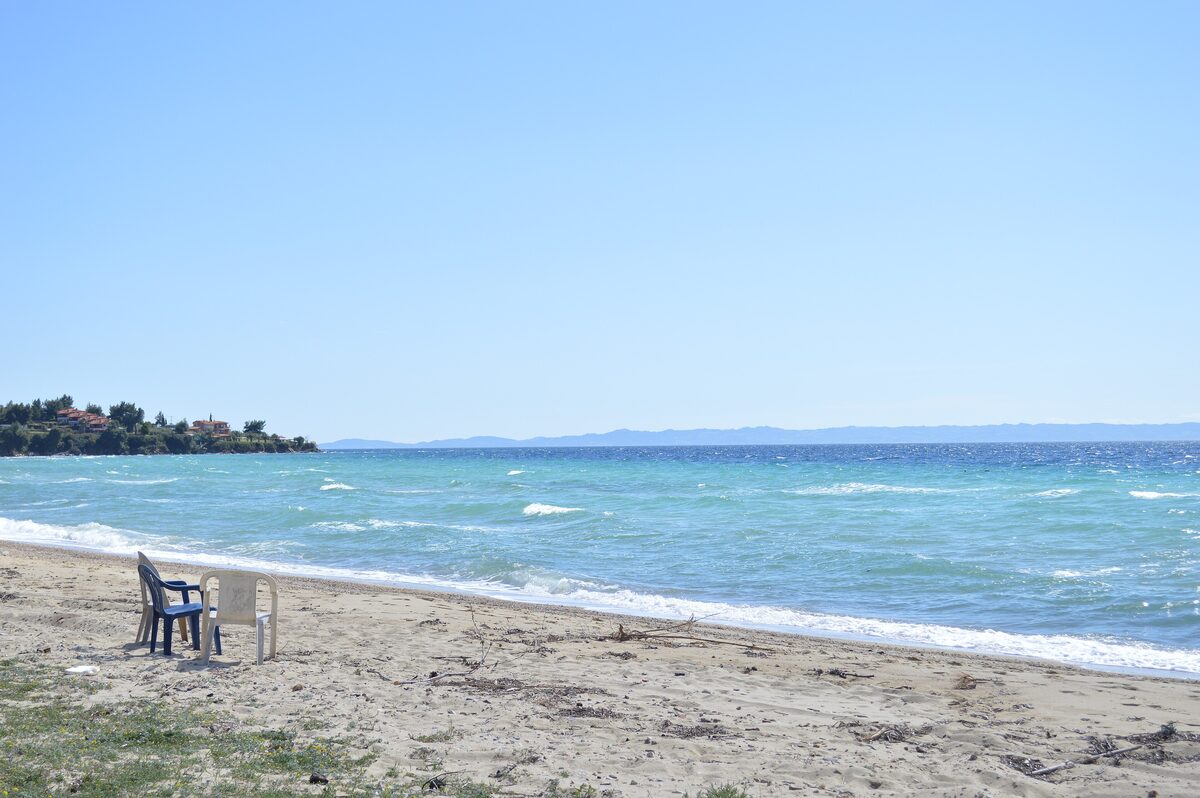 Waves rolling onto the sandy beach at Nikiti under a clear blue sky, with two plastic chairs on the sand and a wooded headland across the bay