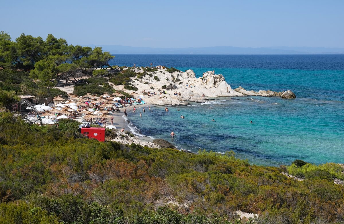 The sandy cove of Orange Beach between pale granite rocks and pines, with umbrellas on the sand and turquoise water