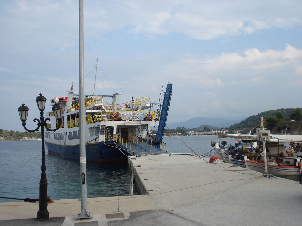 A white passenger ferry tied up at the quay in Ormos Panagias, with a lamp post and boats nearby