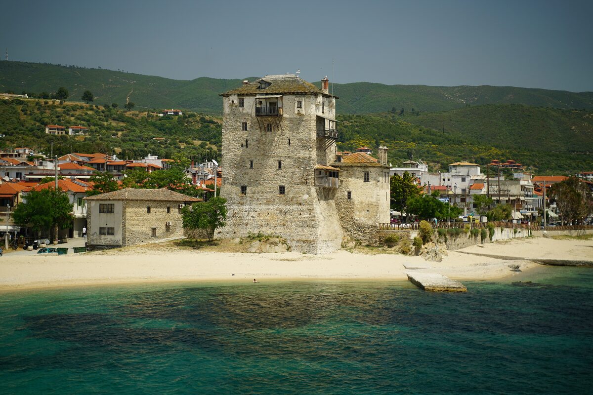 The stone Prosforios Tower on the beach at Ouranoupoli, with houses and hills behind it
