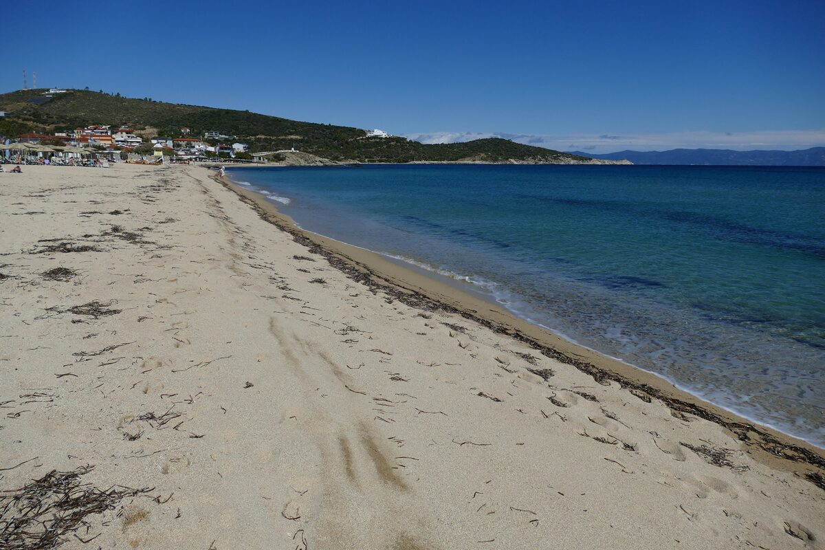 The long, wide sand beach at Sarti stretching away beside clear blue-green water, with the village and green hills at the far end