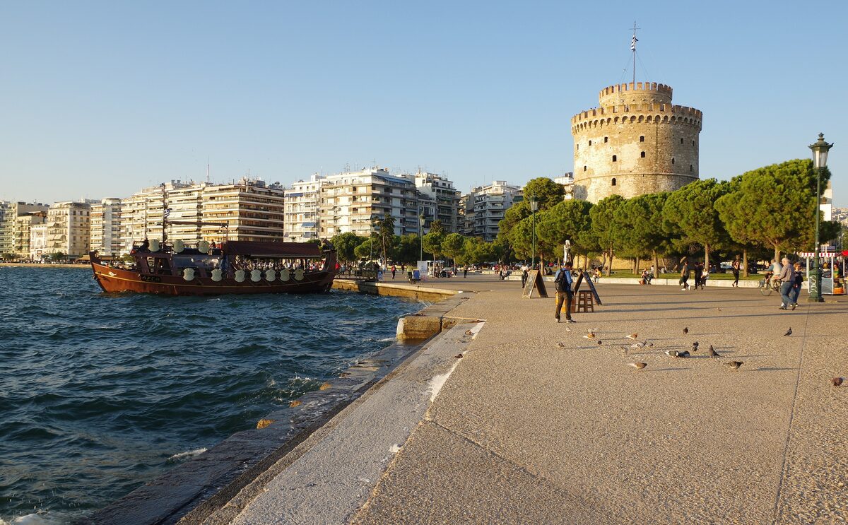 The White Tower of Thessaloniki at the end of the seafront promenade in afternoon light, with apartment blocks and a tour boat along the water