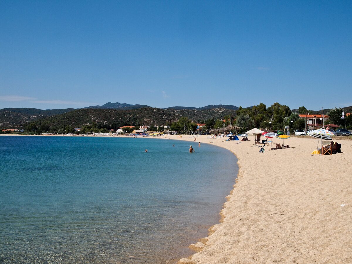 The curve of Toroni's sandy beach beside clear turquoise water, with umbrellas, houses and low hills along the shore