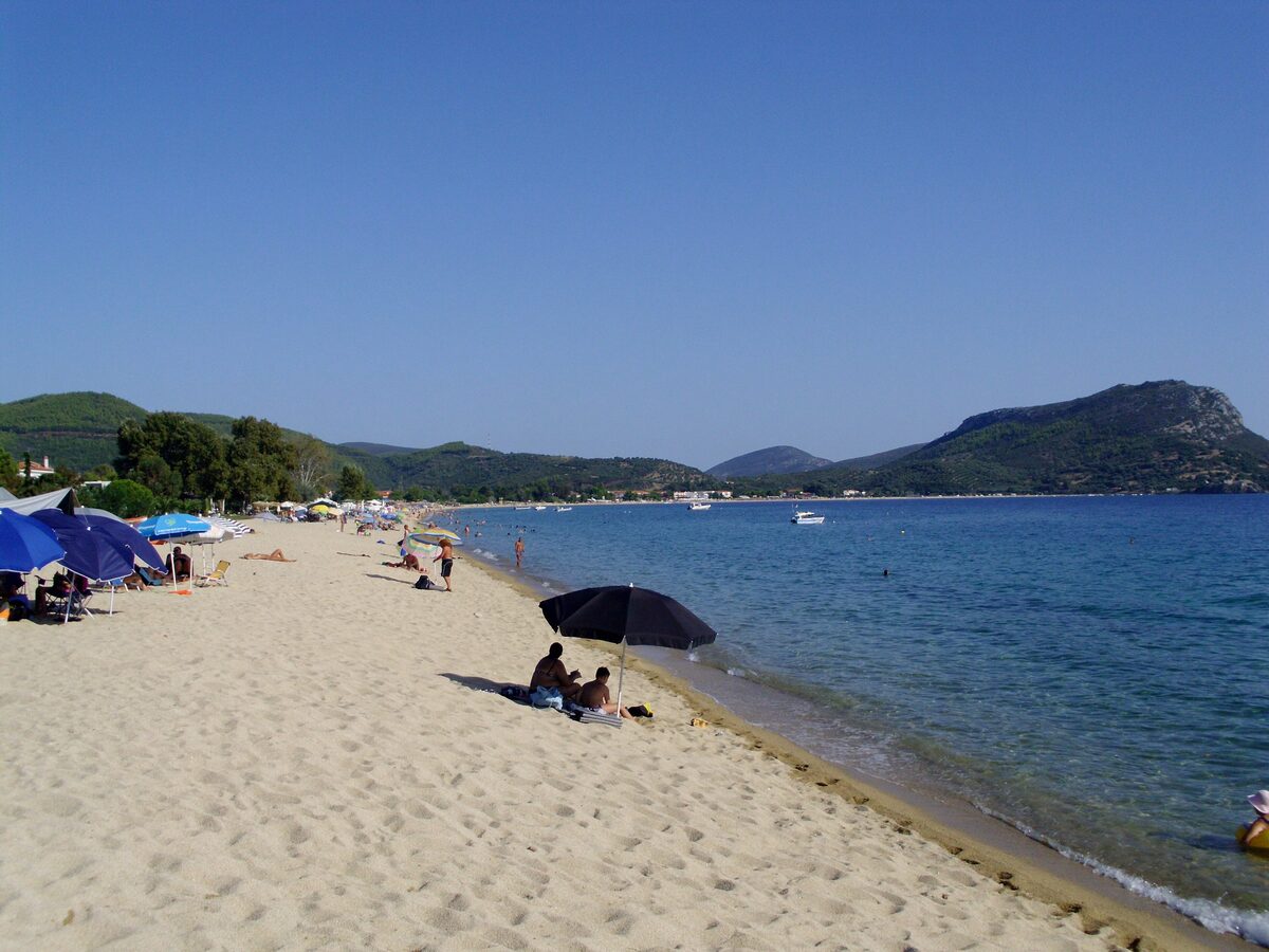 Sunbathers under beach umbrellas on the long sandy beach at Toroni, with calm blue water and a green headland behind
