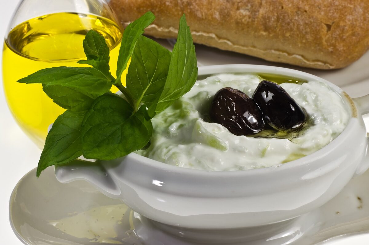 A bowl of tzatziki with mint leaves and black olives, with olive oil and bread beside it