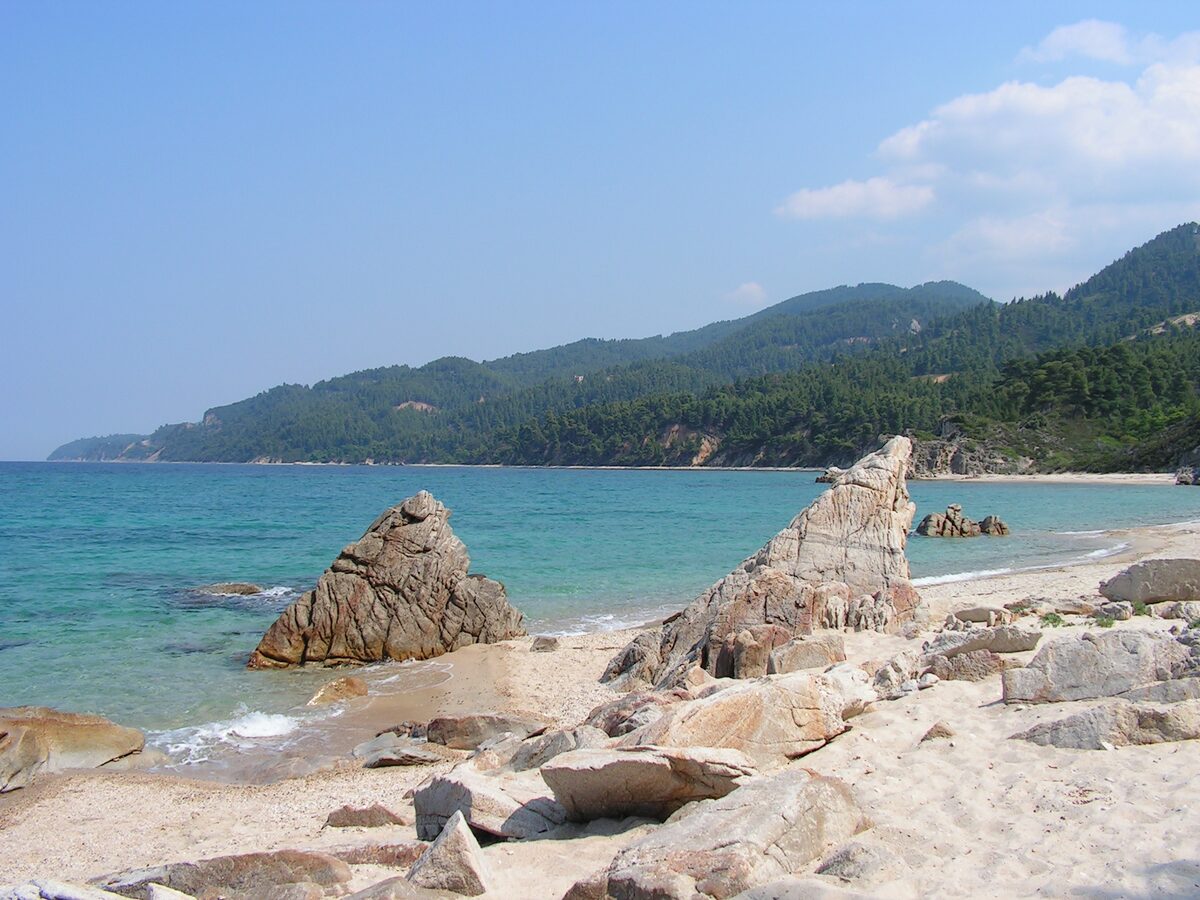 Turquoise water and pale granite rocks on a sandy Vourvourou beach, with forested hills running along the coast behind