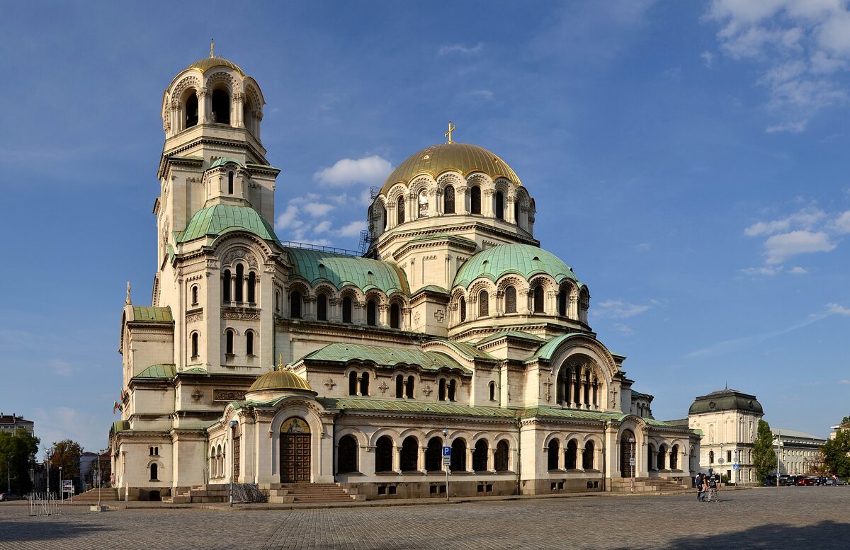 Alexander Nevsky Cathedral with its bell tower, gold dome and green cupolas rising over the wide cobbled square under a blue sky.