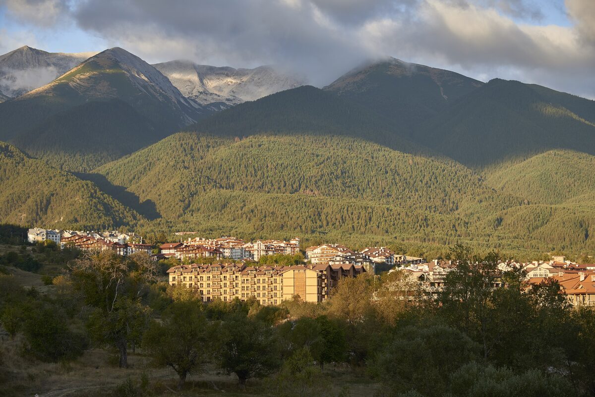 Bansko spread along the foot of the forested Pirin range, with rocky peaks under clouds above the town.