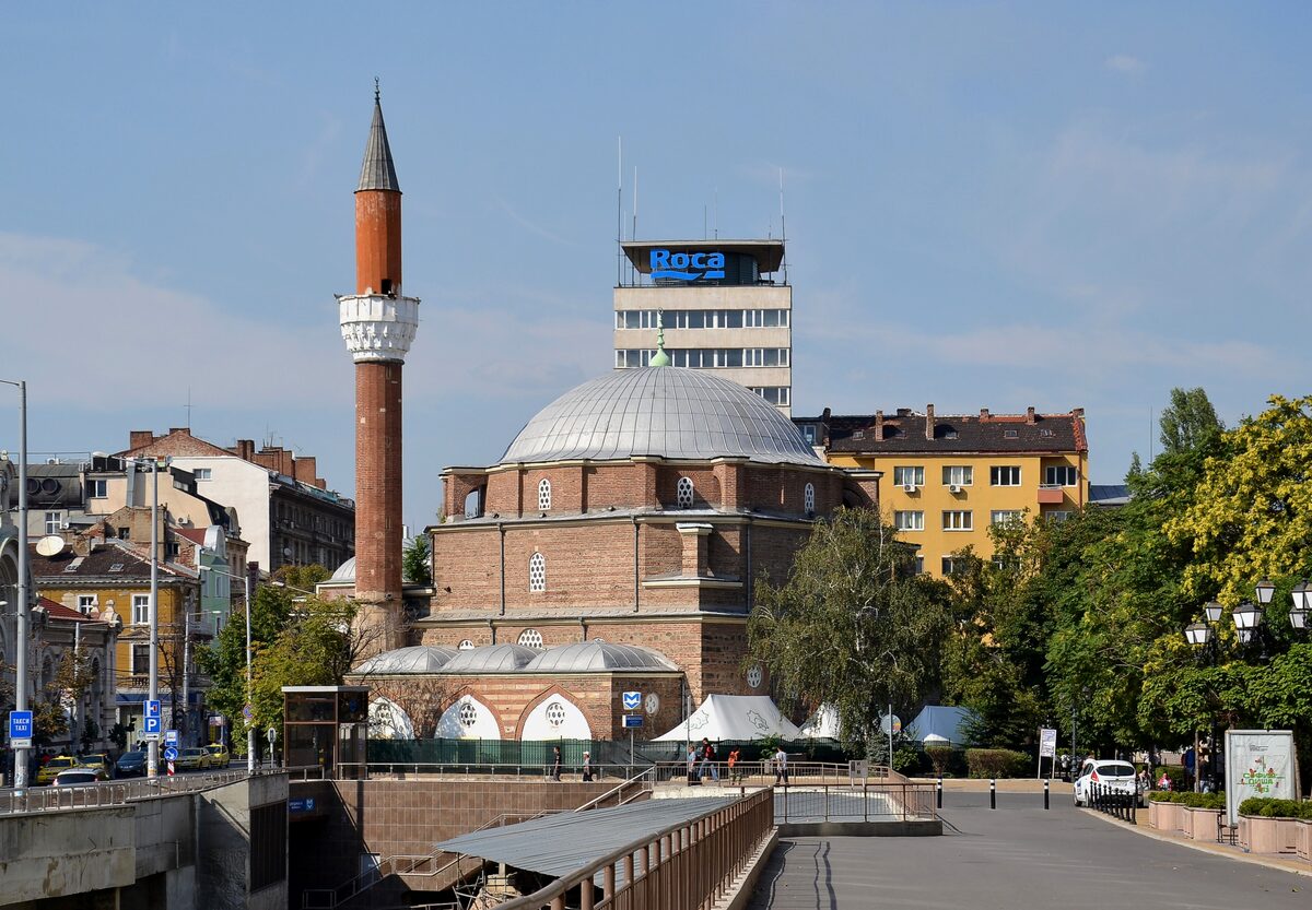 Banya Bashi Mosque with its lead-grey dome and slender brick minaret, seen from the square above the Serdika ruins on a sunny day.