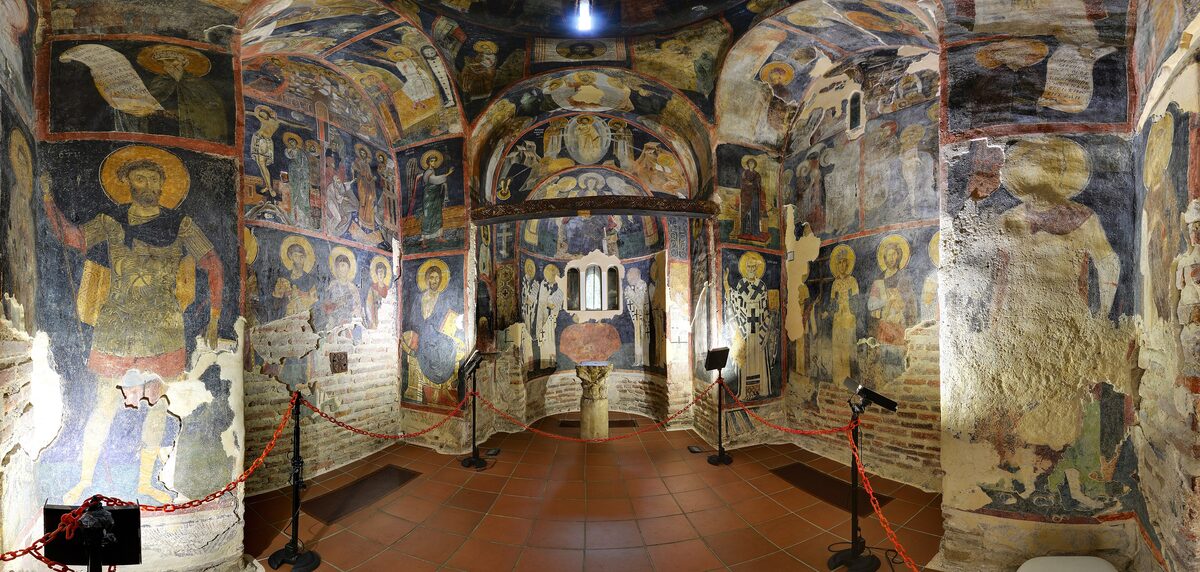 A wide view inside Boyana Church, its small vaulted nave covered floor to ceiling in 13th-century frescoes of saints, behind a red rope barrier.