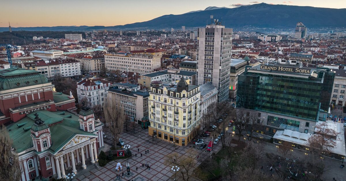 An aerial view of central Sofia at dusk, with the National Theatre, City Garden and Grand Hotel Sofia in front and Vitosha mountain rising behind the rooftops.