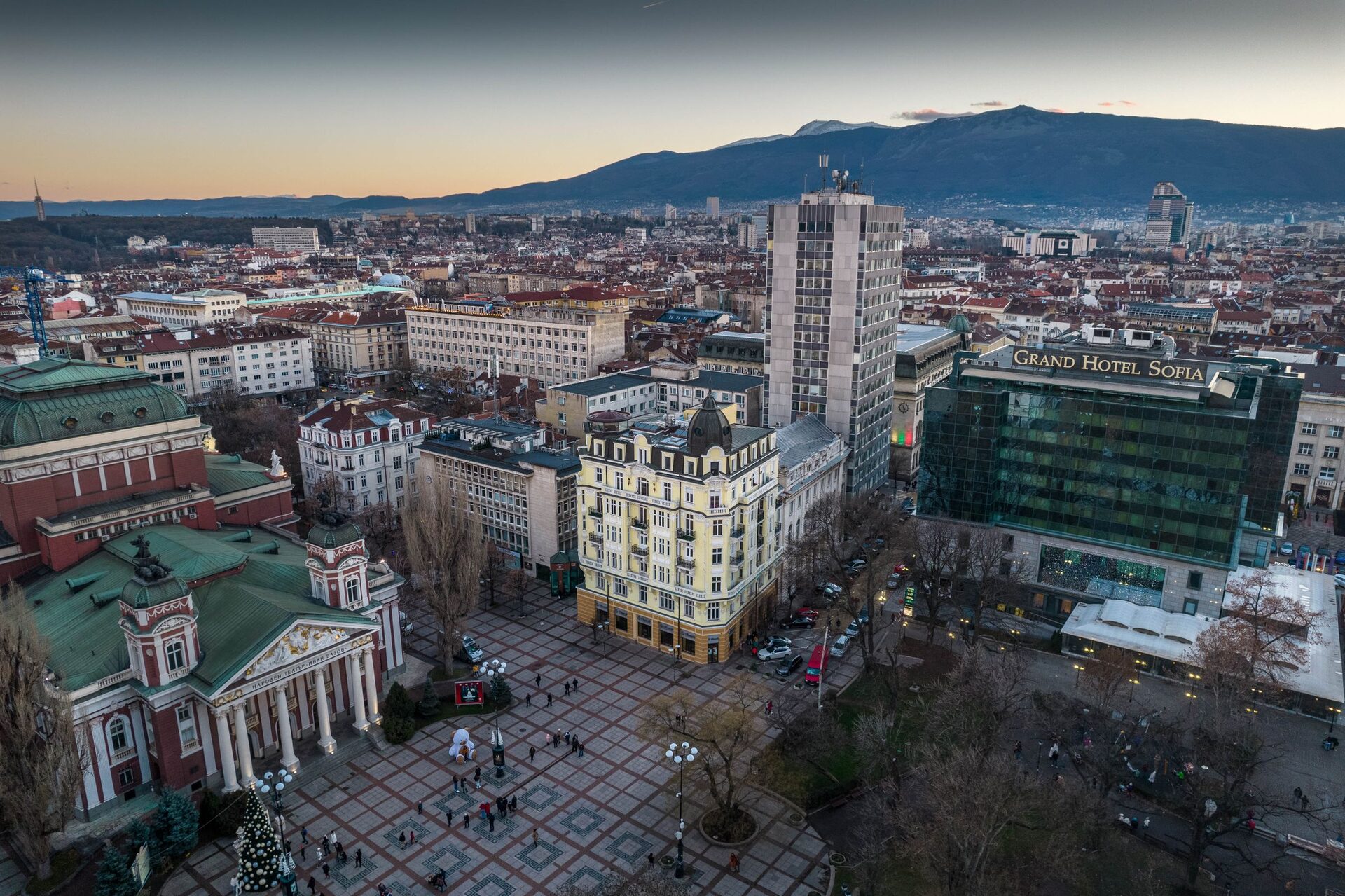 An aerial view of central Sofia at dusk, with the National Theatre, City Garden and Grand Hotel Sofia in front and Vitosha mountain rising behind the rooftops.
