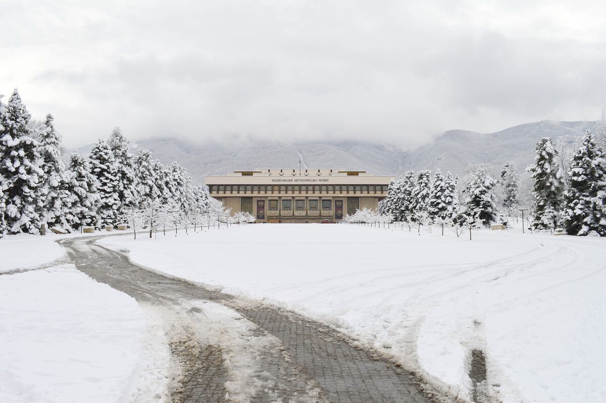 A snowy path through fir trees leading to the National Museum of History in Boyana, with the clouded slopes of Vitosha behind it.