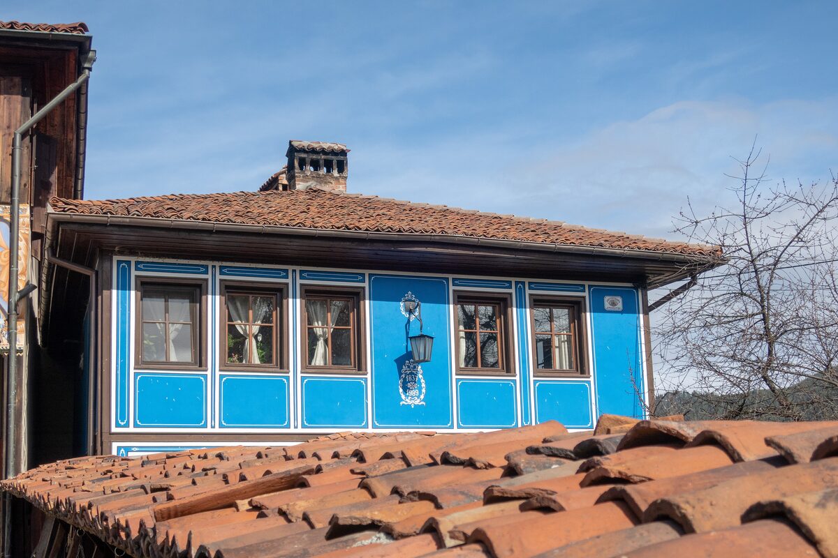 A bright blue National Revival house with white-framed windows and a tiled roof in Koprivshtitsa, seen over old clay roof tiles.