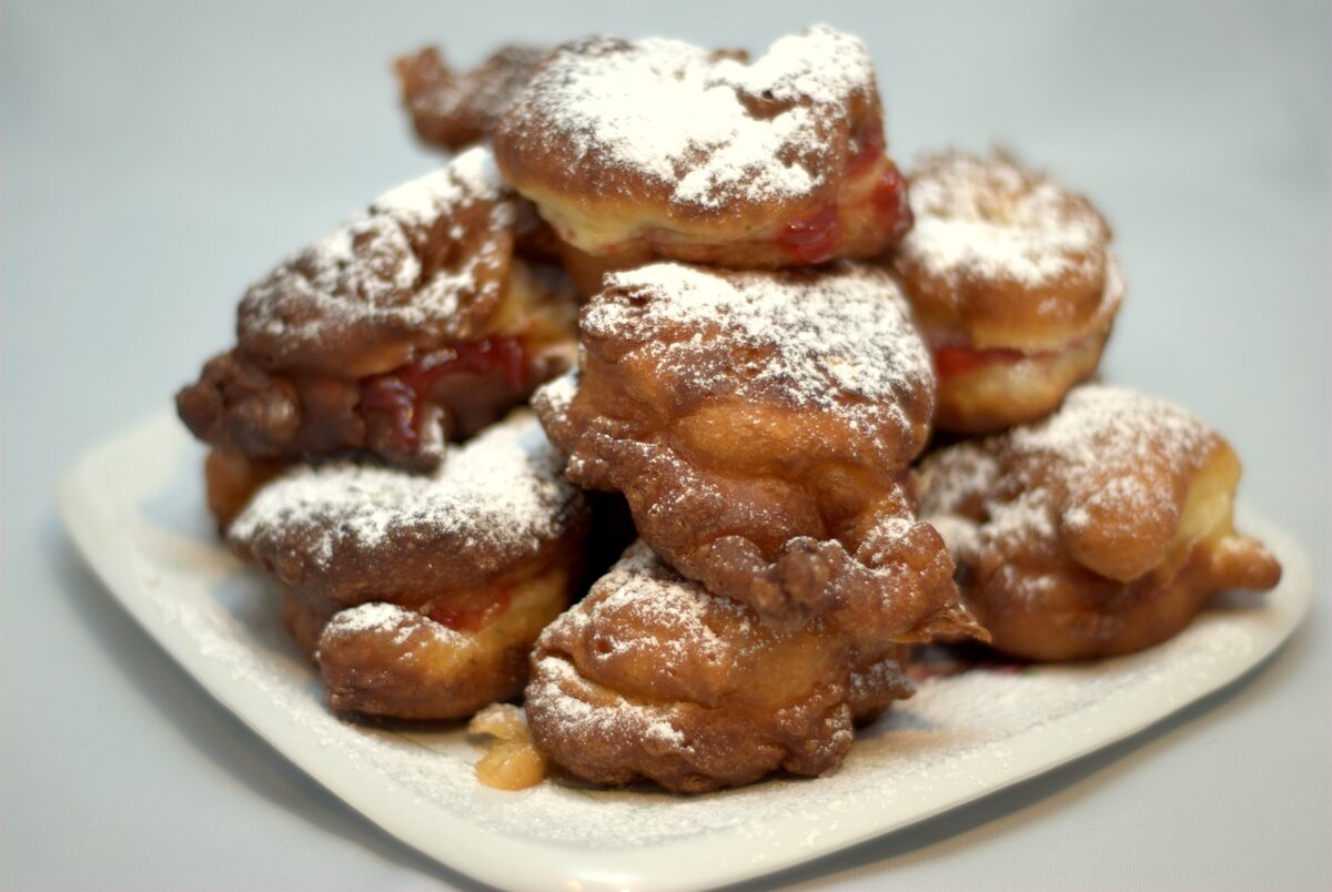 A plate of golden fried mekitsi dusted with powdered sugar, with red jam showing between the pieces.