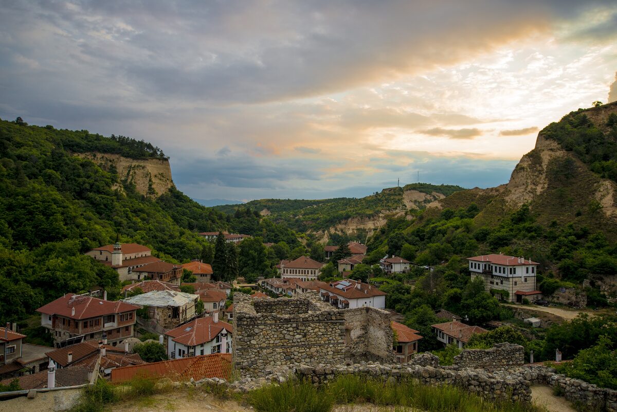 The red-roofed houses of Melnik in a narrow valley between sandstone cliffs and green hills, seen past old stone walls at sunset.