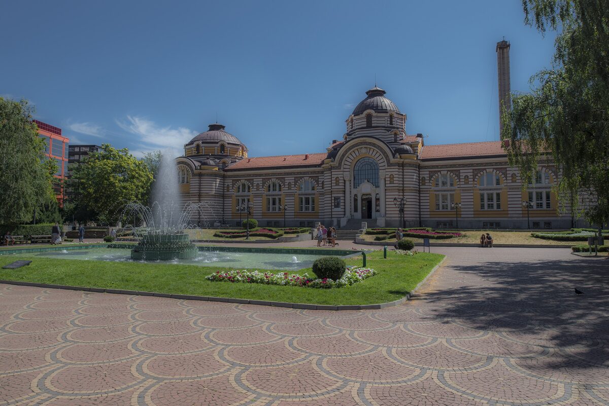 The domed, striped facade of the Central Mineral Baths seen across its garden with a fountain and patterned paving in front.