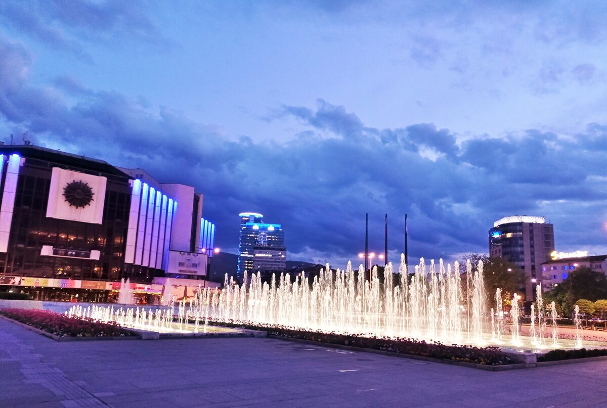 The National Palace of Culture (NDK) at dusk, with its lit facade, the fountains of the plaza in front and towers of the city on the horizon.