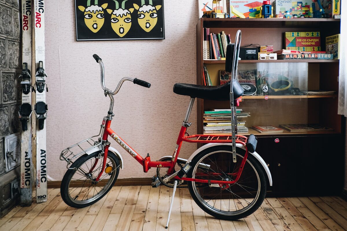 A corner of the Red Flat museum apartment with a red folding bicycle, old skis, a bookcase of Bulgarian books and toys, and a retro poster on the wall.