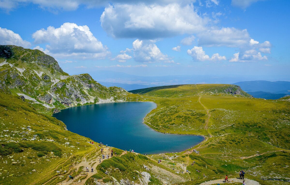 The deep blue Kidney Lake, one of the Seven Rila Lakes, in a green alpine bowl with hikers on the trail along its shore.