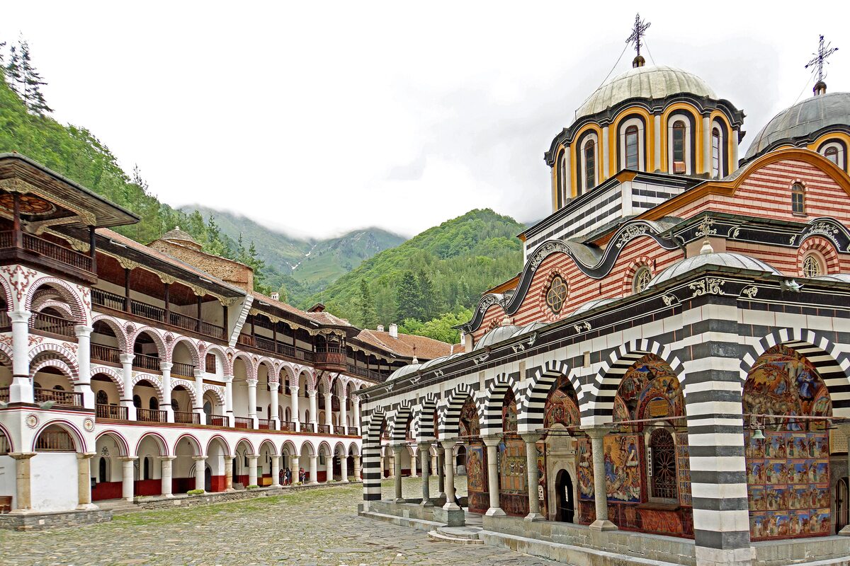 The courtyard of Rila Monastery, with the red-and-white striped arcades and domes of the Nativity church beside the tiers of wooden galleries and forested mountains behind.