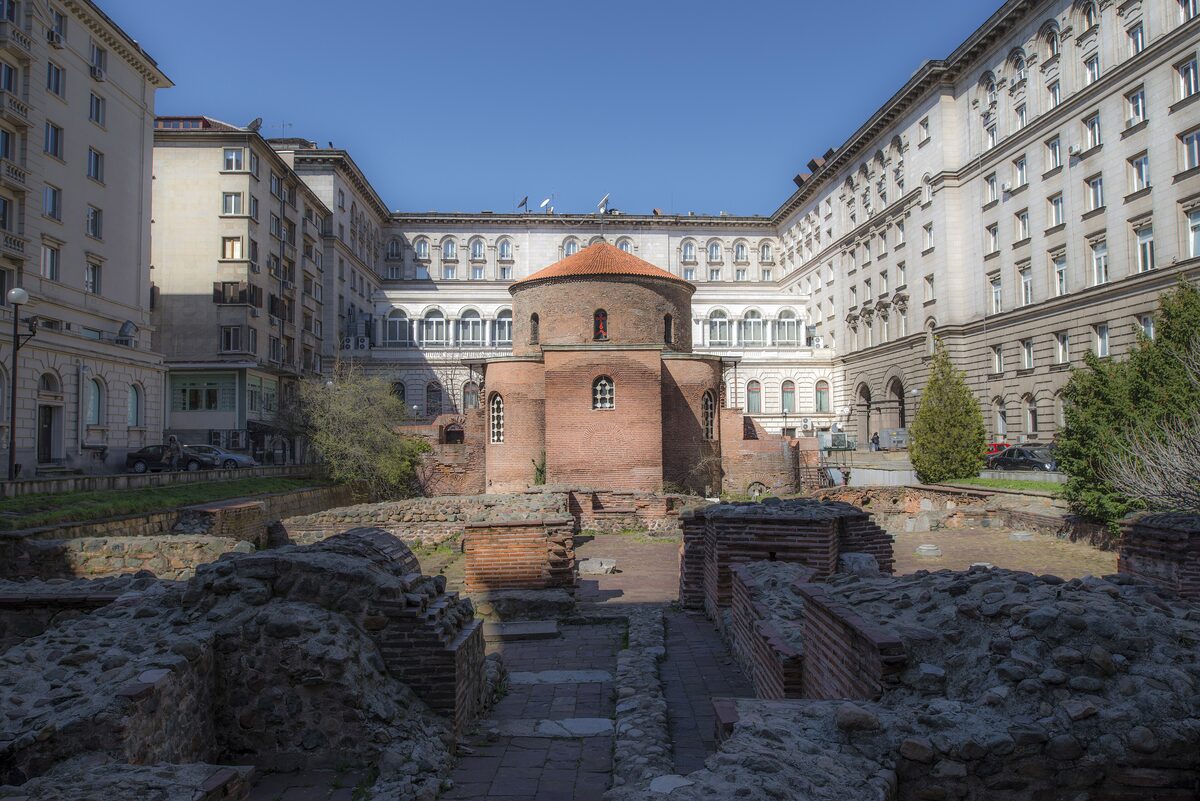 The red-brick Rotunda of St. George standing amid Roman ruins in a courtyard enclosed by the pale stone buildings of the Presidency and the Balkan hotel.