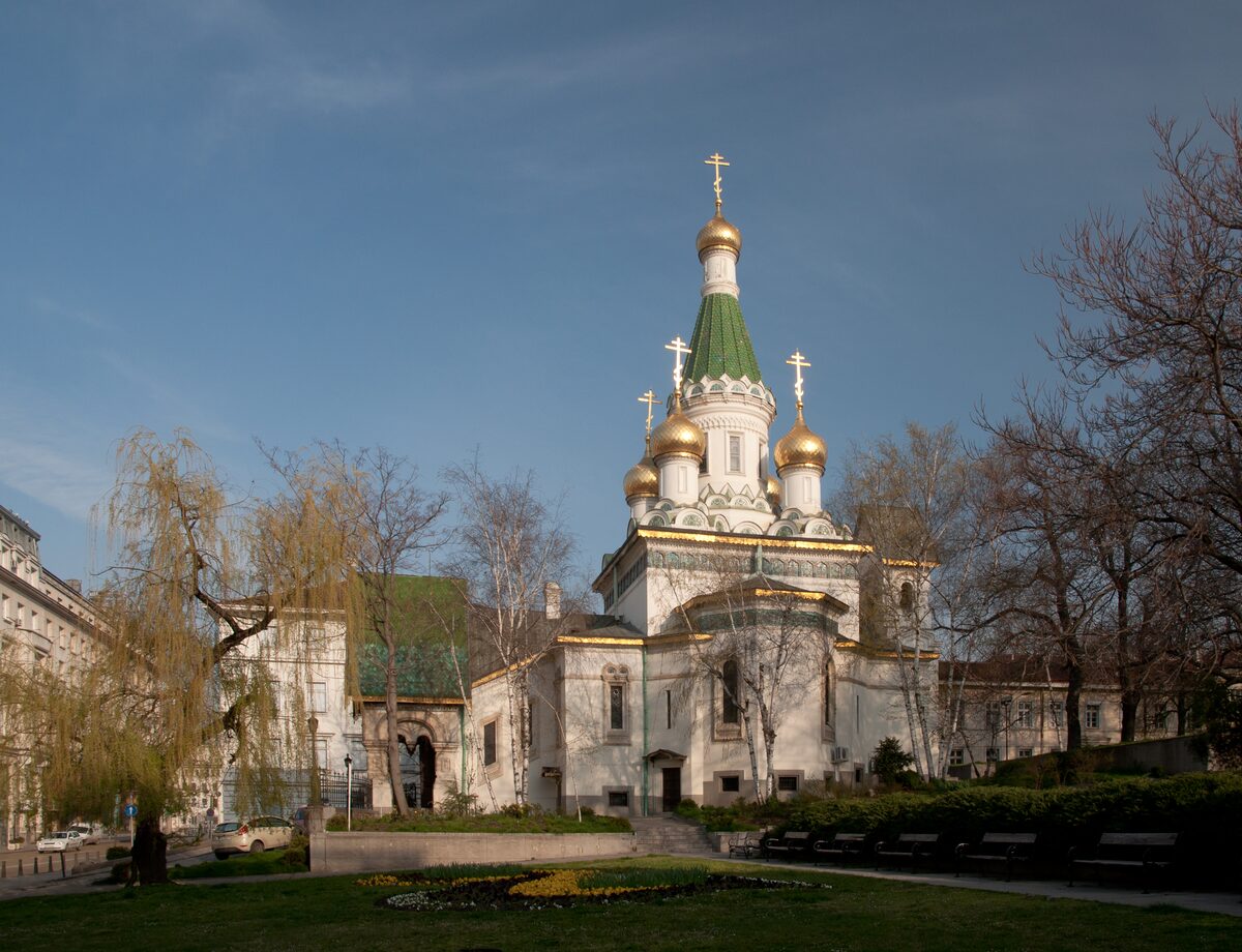 The white Russian Church of St. Nicholas with its green spire and gilded onion domes, framed by willows and bare trees in its small garden.