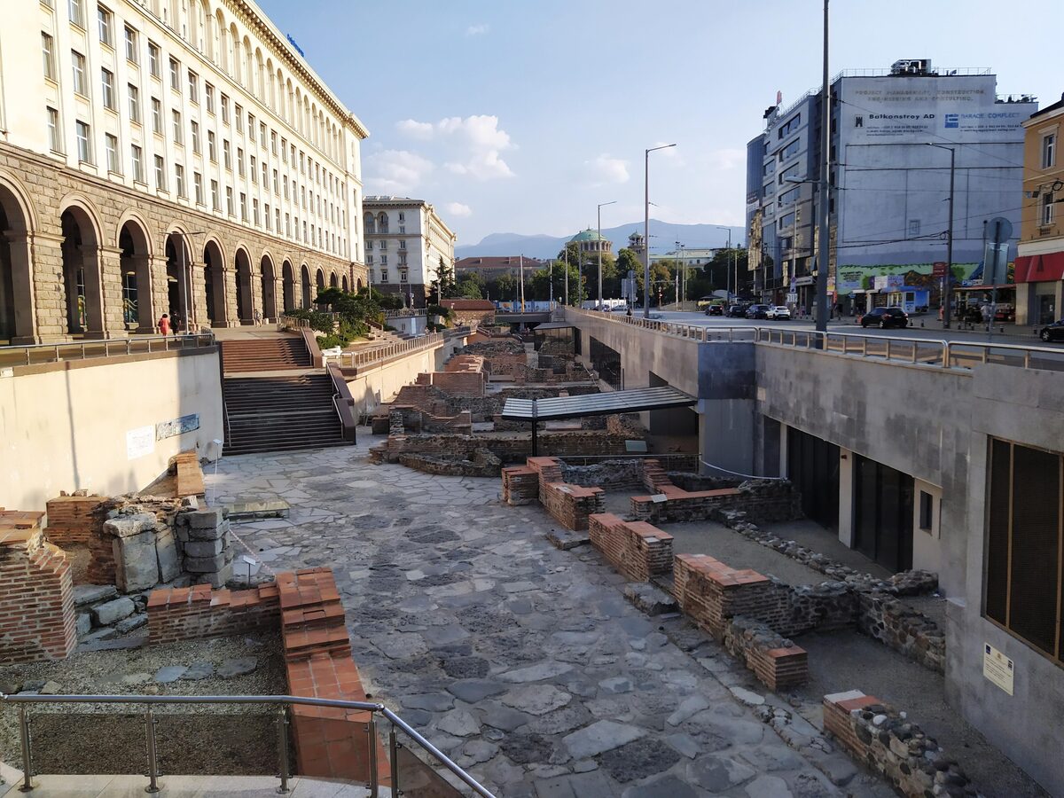 The open-air Ancient Serdica complex at the Largo, with a Roman paved street and restored brick walls below the arcaded socialist-era buildings and Vitosha in the distance.