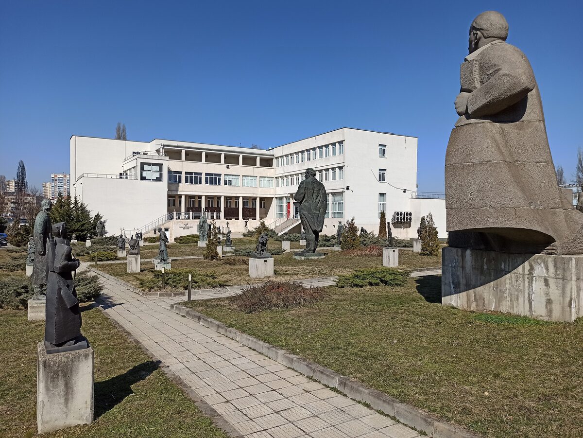 The sculpture park of the Museum of Socialist Art, with a giant stone statue in the foreground and rows of communist-era figures and busts on the lawns before the white museum building.
