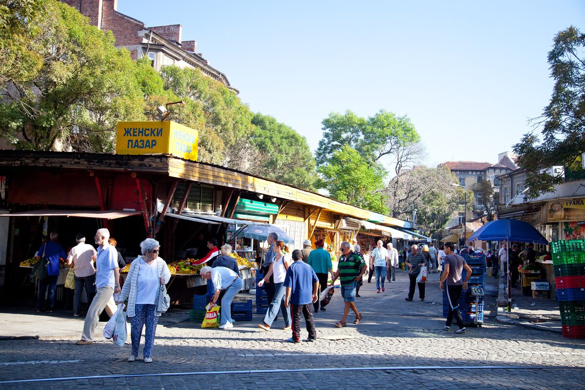 Shoppers walking between the produce stalls of Zhenski Pazar under its yellow 'Женски пазар' sign on a sunny morning.