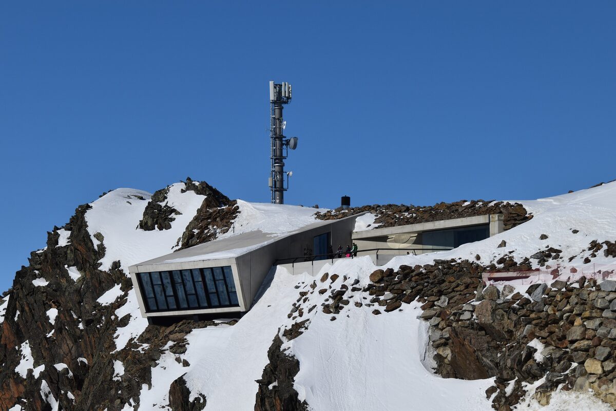 The glass-fronted 007 Elements building on a snowy rock ridge under a deep blue sky, with a mast above it