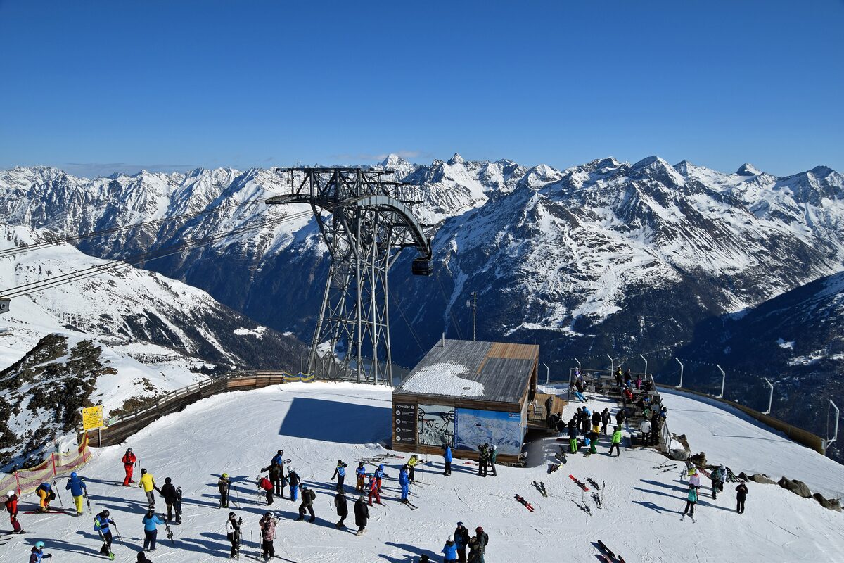 A Gaislachkogl cable car pylon and cabin beside a timber station on a sunny snowy ridge, with skiers gathered in front and mountains behind