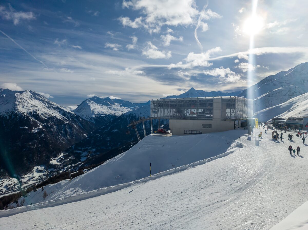 The glass top station of the Giggijoch cable car on a groomed piste, with snowy peaks behind and skiers on the right